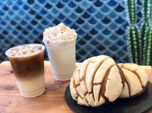 Two drinks, one iced coffee and one creamy beverage topped with whipped cream, alongside two conchas on a wooden table.