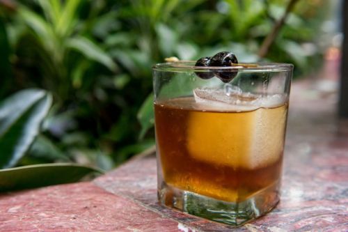 A glass of amber-colored cocktail with ice and a cherry garnish, set against a backdrop of lush green foliage.