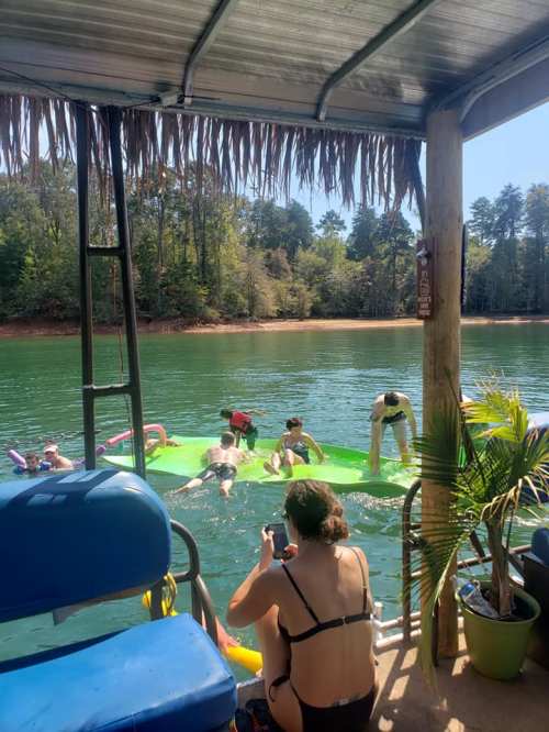 A group of people relaxes on green floats in a calm lake, with trees and a dock in the background on a sunny day.