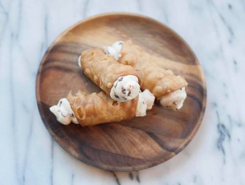 Three cannoli filled with creamy filling on a wooden plate, resting on a marble surface.