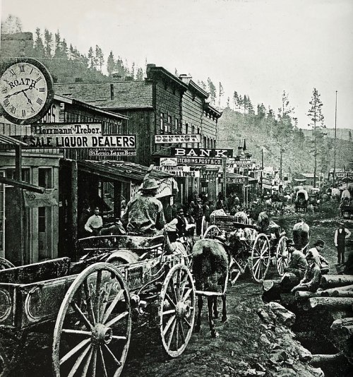 Historic street scene with horse-drawn wagons, storefronts, and people in a mining town. Black and white photo.