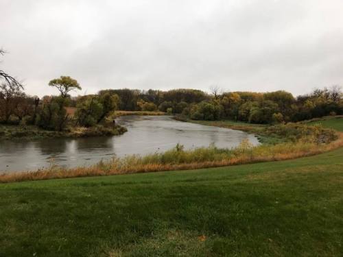 A calm river flows through a landscape of trees and grass under a cloudy sky. Autumn colors are visible in the foliage.