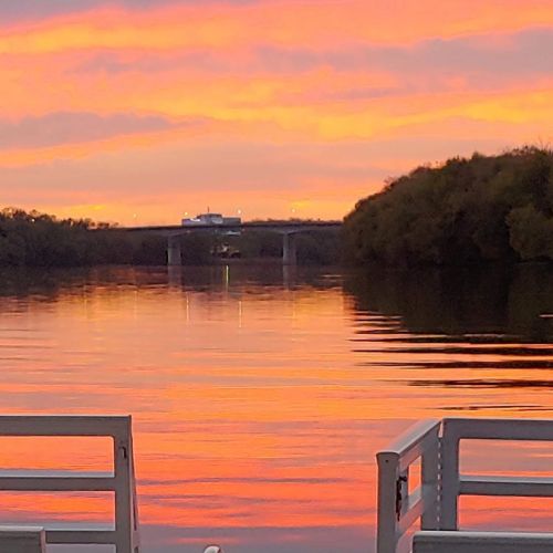 A serene river at sunset, reflecting vibrant orange and pink hues, with a bridge in the distance and trees along the shore.