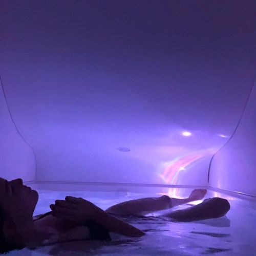 A person relaxes in a dimly lit, purple-tinted floatation tank filled with water.