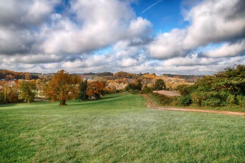 A scenic landscape featuring rolling green hills, colorful autumn trees, and a cloudy blue sky.