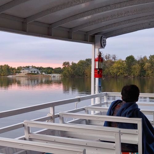 A person sits on a boat, wrapped in a blanket, overlooking calm waters and trees at sunset.