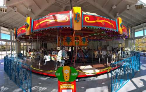 A colorful carousel with white horses, surrounded by a decorative fence, inside a bright, open building.