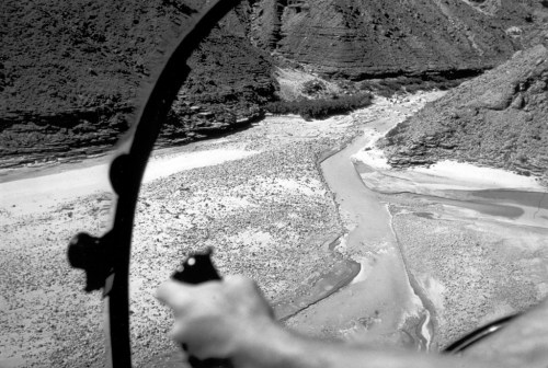 Aerial view of a winding river through rocky terrain, with a hand holding a control stick in the foreground.