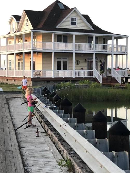 Two children fish from a dock in front of a large, multi-story house at sunset.