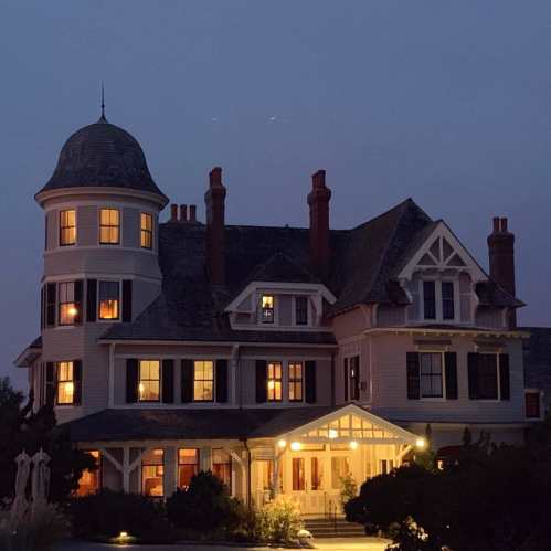 A large, elegant Victorian house at dusk, with illuminated windows and a distinctive turret.