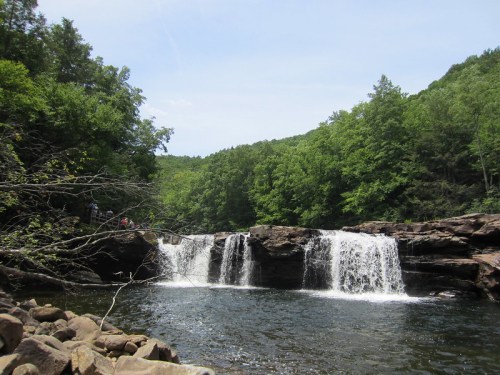 A serene waterfall cascades over rocky ledges, surrounded by lush green trees and a clear blue sky.