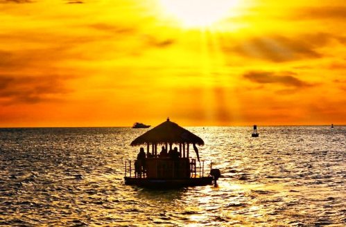A silhouette of a boat with a thatched roof on shimmering water at sunset, with golden rays illuminating the scene.