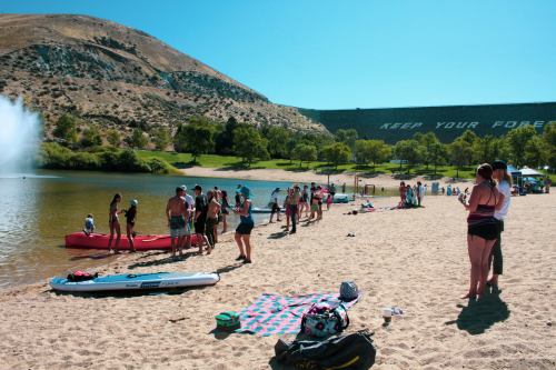 A sandy beach scene with people enjoying water activities, trees in the background, and a hillside with "KEEP YOUR FOREST" sign.