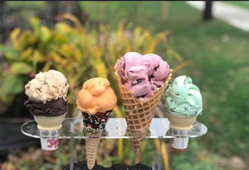 Four ice cream cones in various flavors: chocolate, orange, mint, and pink, displayed on a clear stand outdoors.