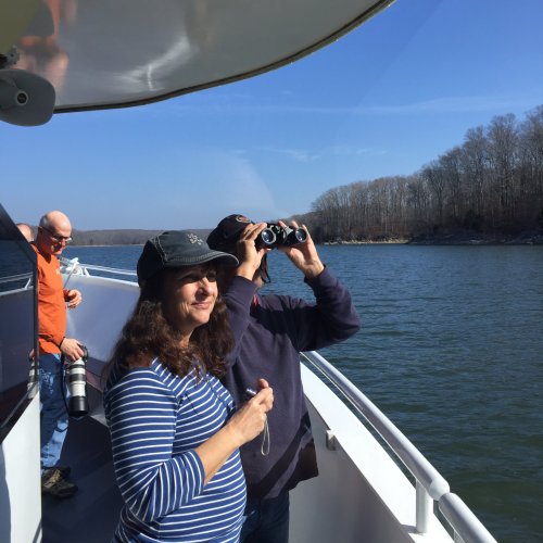 Two people on a boat, one taking photos with a camera, the other enjoying the view, with a scenic river in the background.