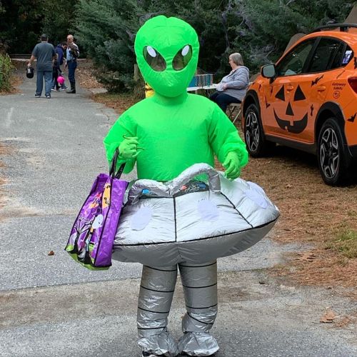 A child in a green alien costume holds trick-or-treat bags while standing next to a parked car decorated for Halloween.