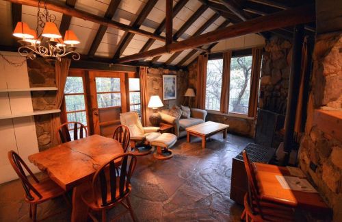 Cozy cabin interior with wooden furniture, stone walls, and large windows overlooking nature. Warm lighting adds charm.