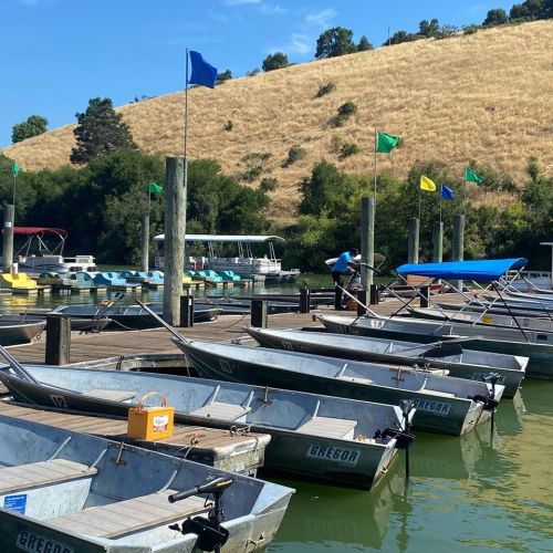 A dock with several small boats tied up, surrounded by green hills and colorful flags fluttering in the breeze.