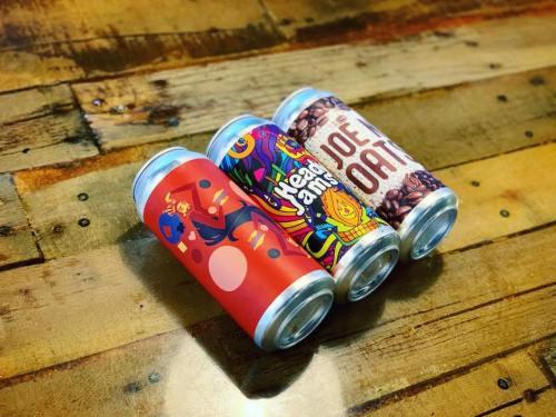 Four colorful beverage cans arranged on a wooden surface, featuring vibrant designs and logos.