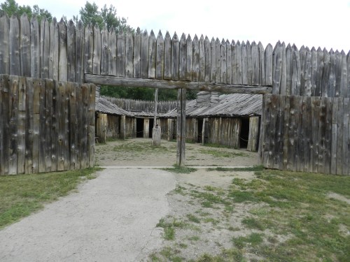 A wooden fort entrance with tall, pointed palisades and a dirt path leading into a grassy area with buildings.
