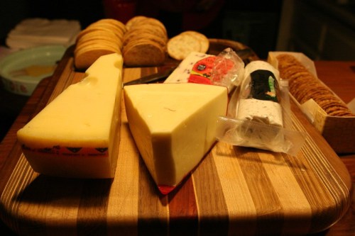 A wooden board displaying various cheeses and crackers, including a wedge of yellow cheese and a log of goat cheese.