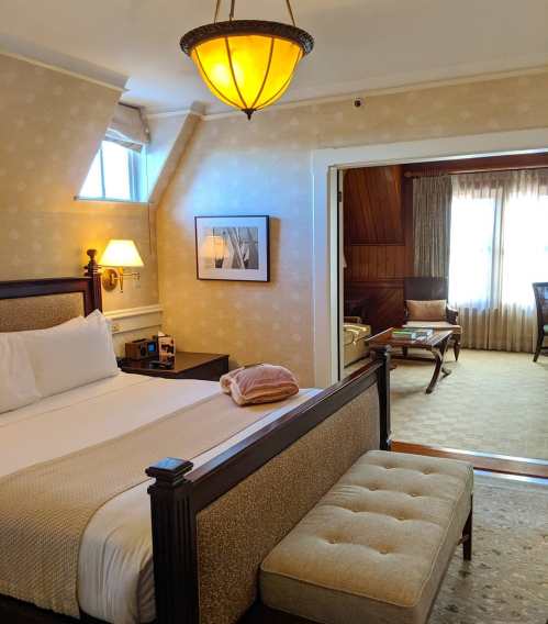 Cozy hotel room featuring a bed, bench, and seating area with natural light from windows. Warm, inviting decor.