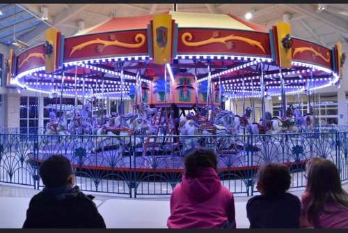 Children watch a brightly lit carousel with colorful horses in an indoor amusement area.
