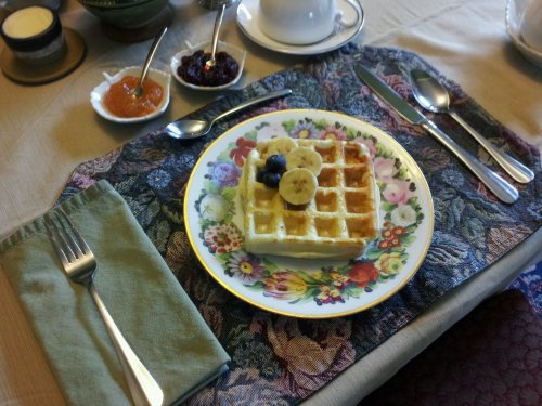 A plate of waffles topped with banana and blueberries, surrounded by small bowls of jam and a cup on a floral tablecloth.