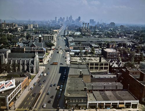 Aerial view of a cityscape with streets, buildings, and distant skyline under a cloudy sky.