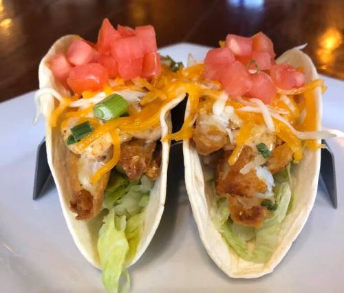 Two crispy fish tacos topped with shredded cheese, diced tomatoes, and lettuce, served on a white plate.