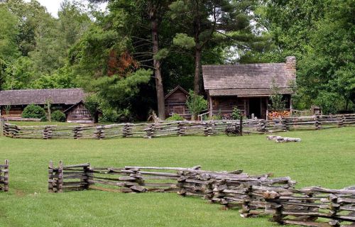 A rustic scene featuring log cabins surrounded by a wooden fence and lush greenery.