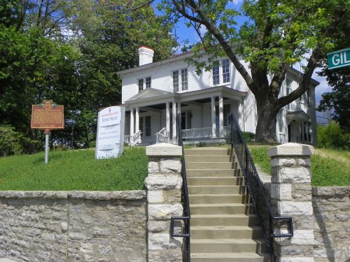 Historic white house with a porch, surrounded by greenery, featuring a sign and steps leading up to it.