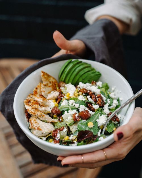 A person holds a bowl of salad with chicken, avocado, corn, nuts, and cheese, served on a wooden table.