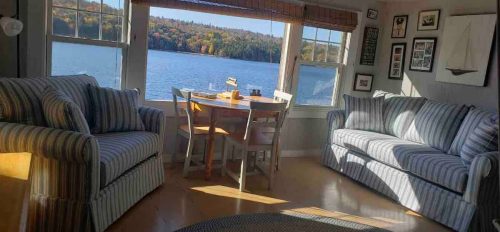 Cozy living area with striped sofas, a dining table, and a view of a lake surrounded by autumn trees.
