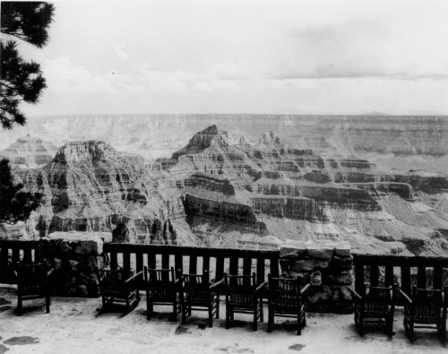Black and white photo of the Grand Canyon, featuring wooden chairs overlooking the vast landscape.