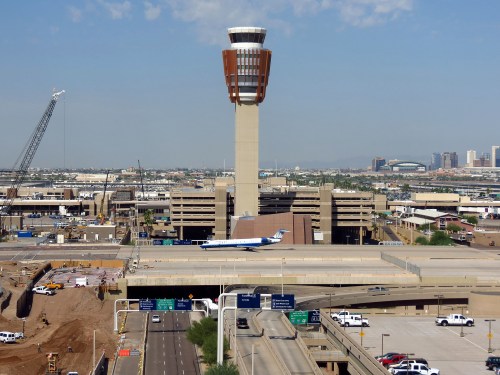Aerial view of an airport with a control tower, parking structure, and a plane on the runway, surrounded by cityscape.