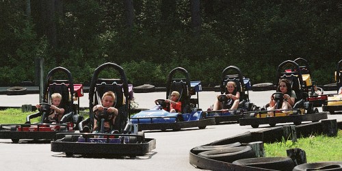 A group of children racing go-karts on a track surrounded by trees and tires.