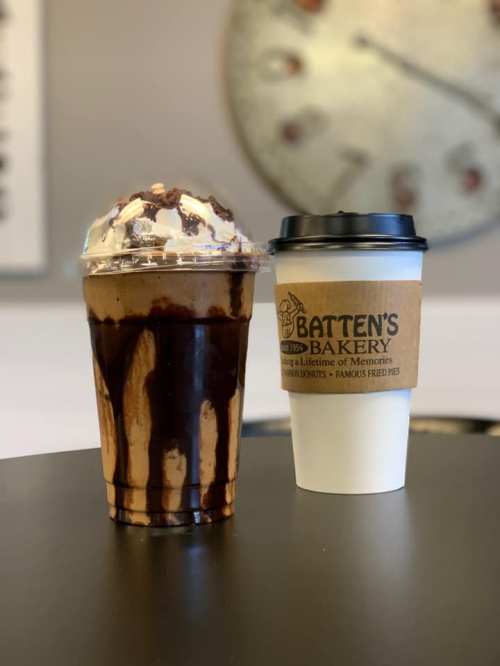 A chocolate drink topped with whipped cream and a coffee cup from Batten's Bakery on a table.