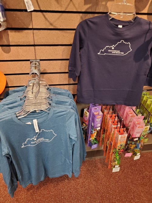 Two long-sleeve shirts in blue and navy, featuring a Kentucky outline design, displayed on a rack with colorful accessories.