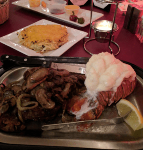 A plate with grilled steak topped with mushrooms and onions, a lobster tail with butter, and a lemon wedge.