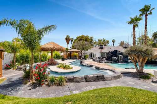 A sunny backyard with a pool, waterfall, palm trees, and a thatched gazebo surrounded by lush greenery and flowers.