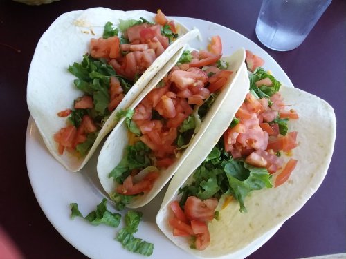 Three soft tacos filled with lettuce, diced tomatoes, and cheese on a white plate, with a glass of water in the background.