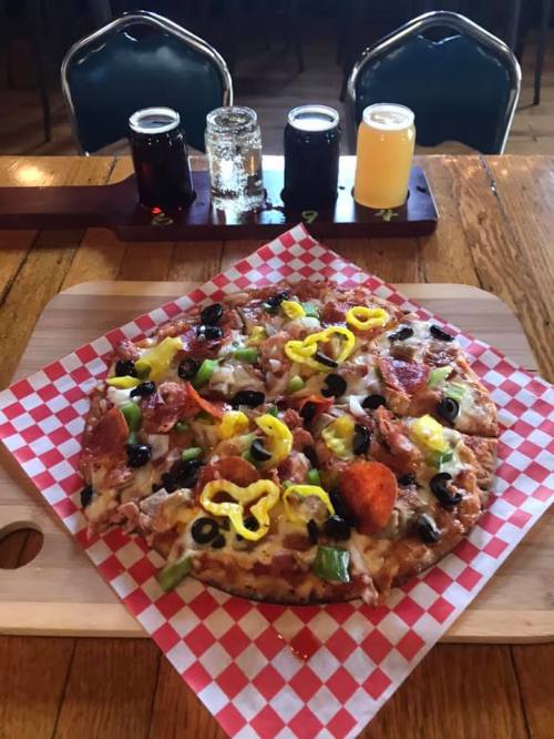 A pizza topped with pepperoni, olives, green peppers, and yellow peppers, served on a wooden board with drinks in the background.