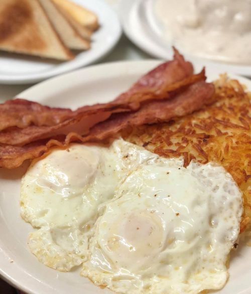 A plate of breakfast featuring two fried eggs, crispy bacon, and golden hash browns, with toast in the background.