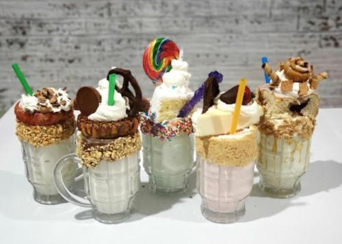 A variety of colorful milkshakes topped with whipped cream, candies, and cookies in glass mugs on a white table.