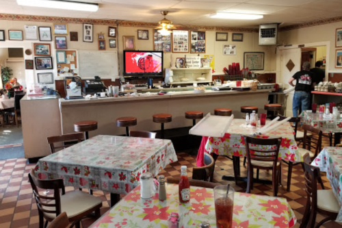 A cozy diner with floral tablecloths, a counter, and a TV, featuring a casual atmosphere and a staff member in the background.