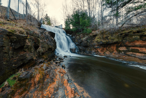 A serene waterfall cascades over rocky terrain into a calm pool, surrounded by trees and a fence in the background.