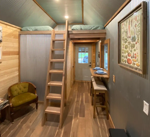 Cozy tiny home interior with a lofted bed, wooden ladder, and rustic decor, featuring a small dining area and door.