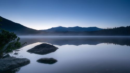 A serene lake at dawn, with mist rising and mountains silhouetted against a blue sky. Rocks are visible in the foreground.