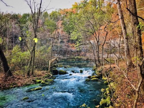 A serene river flows through a forest with autumn foliage, surrounded by trees and rocky banks.